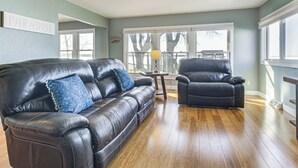 A living room with a black leather sofa, a black leather armchair, and a wooden coffee table. There is a window with a view of trees and a deck outside. - Shawano Lake House w/ Boat Dock, Kayaks, & Grill! (Cecil)