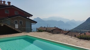 A swimming pool with a clear blue water surface, surrounded by a stone deck and a metal fence. In the background, there are houses with red and beige facades, and a mountain range is visible. - BLONDIE HOUSE (Fonteno)