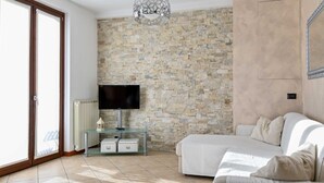 Un salon moderne avec un mur en pierre, un téléviseur à écran plat, une table basse en verre, un canapé blanc et une porte-fenêtre coulissante donnant sur un espace extérieur.