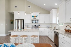 A modern kitchen with white cabinetry, a central island with stools, stainless steel appliances, and pendant lighting. - Two Adjacent Cabins, Sleeps 18 (Bolivar Peninsula)