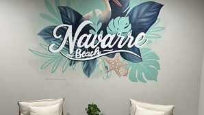 A room with two wooden chairs, a small black table with a plant, and a wall mural reading "Navarre Beach". - Charming 3-bedroom house with WiFi, AC in fabulous Navarre Beach 7 min away. (Navarre)