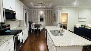 A modern kitchen with white cabinetry, a granite countertop, and a central island. There is a dining area with a table and chairs, and a view into the living room. - Charming 3-bedroom house with WiFi, AC in fabulous Navarre Beach 7 min away. (Navarre)
