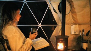 Une personne est assise dans une tente, en train de lire un livre, et un poêle à bois se trouve à proximité.