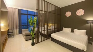 A modern hotel room with a bed, a desk, a chair, a potted plant, and a view of the cityscape. - The Pines by Amanjiwa (Melaka)