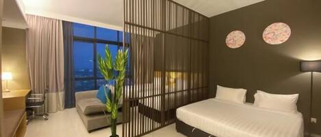 A modern hotel room with a bed, a desk, a chair, a potted plant, and a view of the cityscape.