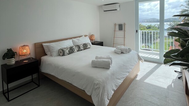 A modern bedroom with a large bed, a nightstand with a lamp, and a view of the outdoors through a sliding glass door. - Private Island 1BR Condo with Oceanview, Pool View, 500MB WiFi & A/C (Fajardo)