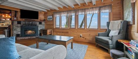 Acogedor interior de cabaña con chimenea de piedra, paredes de madera y amplias ventanas que dan a un paisaje exterior.
