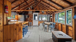 L’intérieur d’un chalet rustique, avec un plafond en bois, un coin cuisine, un espace repas et de grandes fenêtres.