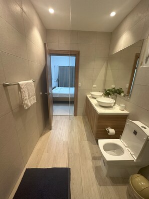 Une salle de bain moderne équipée de toilettes, d’un bidet, d’un lavabo et d’un miroir. Un porte-serviettes se trouve à gauche, et une porte donne accès à une chambre.