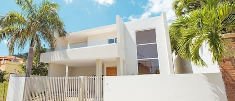 A modern two-story house with a balcony, surrounded by palm trees and a white fence.