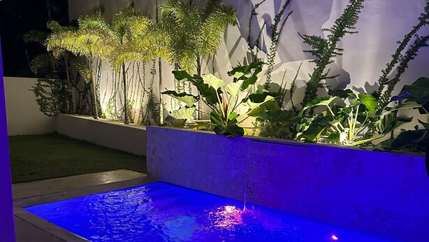 Ein moderner Poolbereich im Freien mit einem blau beleuchteten Wasserspiel, umgeben von üppigem Grün und einer weißen Mauer.