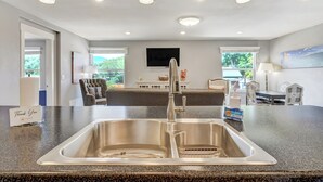 A modern kitchen with a stainless steel sink, overhead pendant lights, and a flat-screen TV mounted above the counter.
