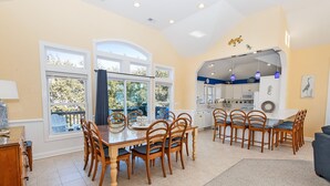 Uma sala de jantar espaçosa com uma grande mesa de madeira e cadeiras, uma cozinha ao fundo, e janelas amplas com vista para o exterior.