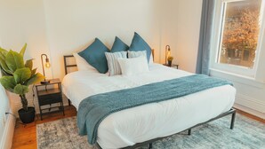 A bedroom with a bed, pillows, a bedside table, a lamp, a plant, and a window. - Large Condo in the heart of GR (Grand Rapids)