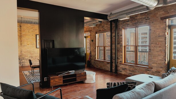 A modern living room with a large flat-screen TV, a sofa, a coffee table, and a brick wall. - Large Condo in the heart of GR (Grand Rapids)