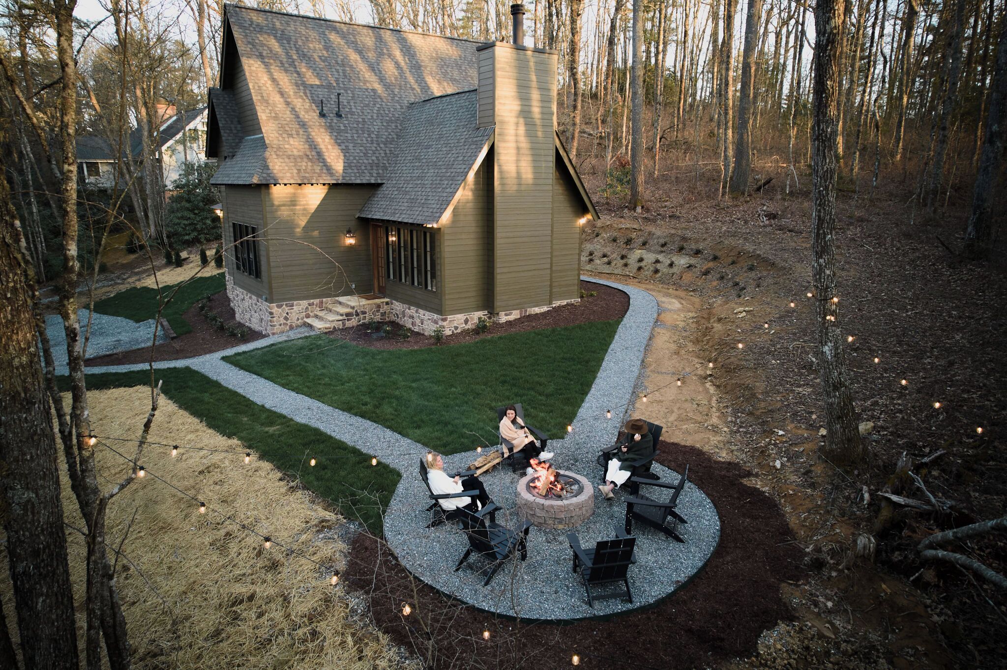 Une maison avec des fondations en pierre et un toit en pente, entourée d’une pelouse bien entretenue et d’un espace barbecue avec des sièges d’extérieur.