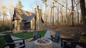 Een blokhut met een vuurkuil van steen, omringd door Adirondack-stoelen, gelegen in een bebost gebied.
