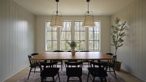 Een eetkamer met een houten tafel, zwarte stoelen, twee hanglampen en een groot raam met uitzicht op bomen.