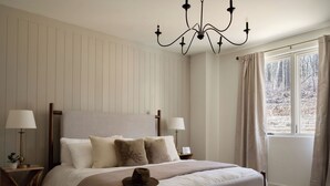 Een slaapkamer met een groot bed, twee nachtkastjes, een kroonluchter, een raam met gordijnen en uitzicht op bomen.