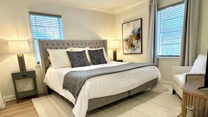 A bedroom with a bed, two bedside lamps, a ceiling fan, a window with blinds, a painting on the wall, and a chair.
