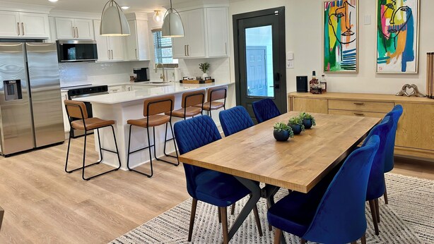 A modern kitchen with white cabinets, stainless steel appliances, and a central island with bar stools. There are two paintings on the wall, a wooden dining table with blue chairs, and a patterned rug on the floor.