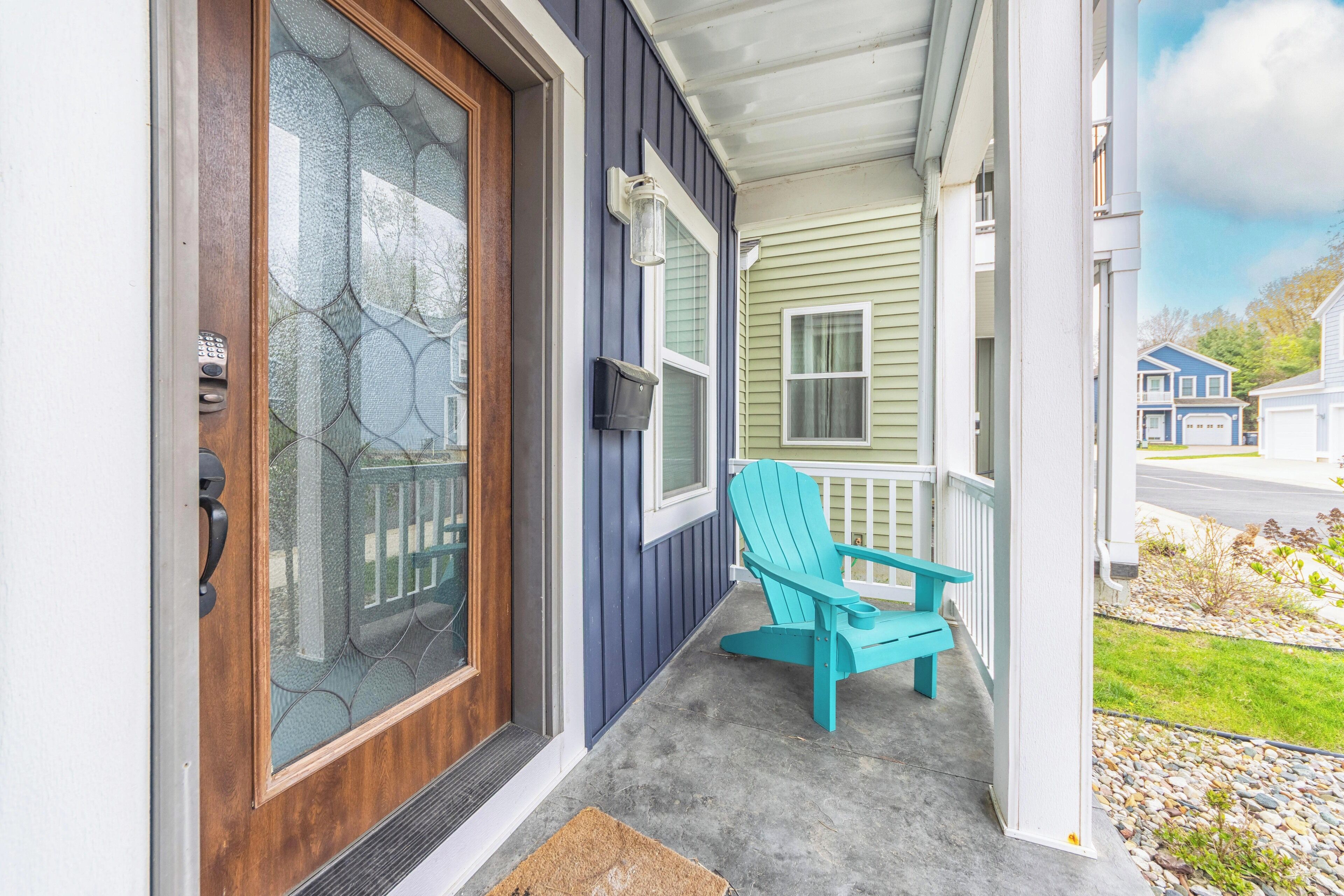 Une véranda avec un fauteuil Adirondack de couleur turquoise, une porte en bois dotée d’un panneau de verre et une boîte aux lettres fixée au mur.