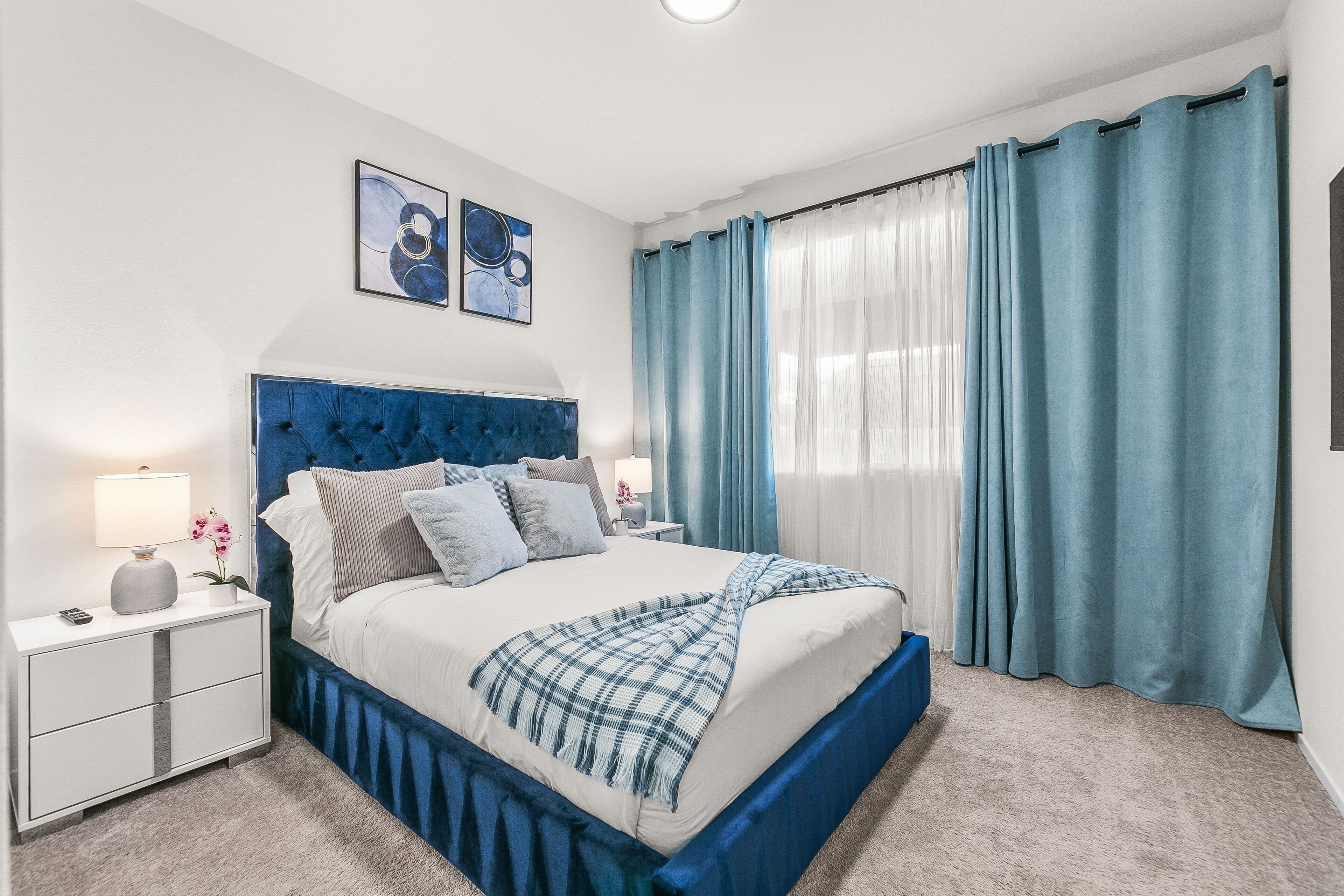 Ein modernes Schlafzimmer mit einer gepolsterten Kopfstütze, einem Bett mit weißer Bettwäsche und blauen Akzenten, einem Nachttisch mit Lampe und Blick ins Freie durch transparente Vorhänge.
