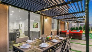Un espace de restauration en plein air agrémenté d’une pergola, d’une table à manger dressée avec de l’assiette blanche, des verres et des couverts, ainsi qu’un coin salon avec des coussins rouges.