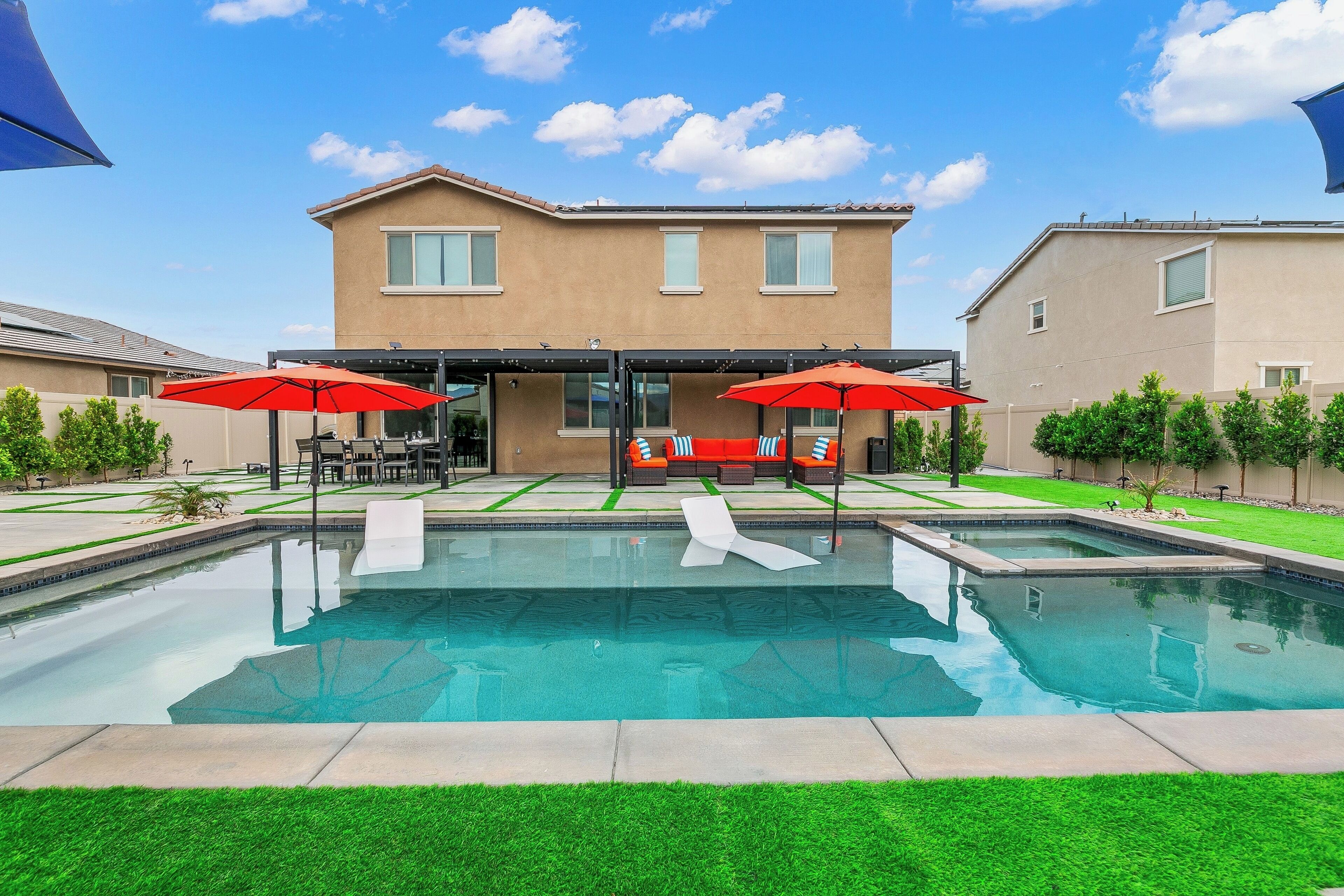 Una piscina con dos sombrillas rojas, un patio con muebles de exterior y una casa de dos pisos al fondo.