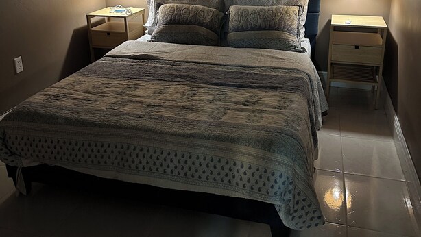 Una cama bien tendida con una colcha estampada y almohadas, flanqueada por dos mesitas de noche con lámparas, en una habitación con paredes lisas y suelo de baldosas.