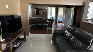 Una sala de estar con televisor, sofá de cuero y comedor, con una cocina al fondo.