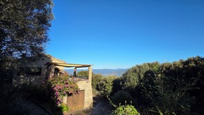 Un edificio de piedra con una pérgola de madera, rodeado de exuberante vegetación y un cielo azul despejado.