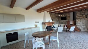 Una cocina con armarios blancos, una mesa de comedor de madera y sillas blancas. Se observa una pared de piedra y una escalera de madera al fondo.