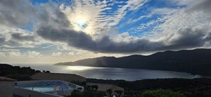 Una piscina con lettini, un paesaggio montuoso e un corpo d'acqua sotto un cielo parzialmente nuvoloso.