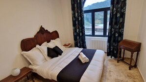 Una cama bien hecha con ropa de cama blanca y negra, un cabecero de madera y una mesita de noche con una lámpara. Se ve una ventana con cortinas, y una pequeña mesa de madera está al lado de la cama.