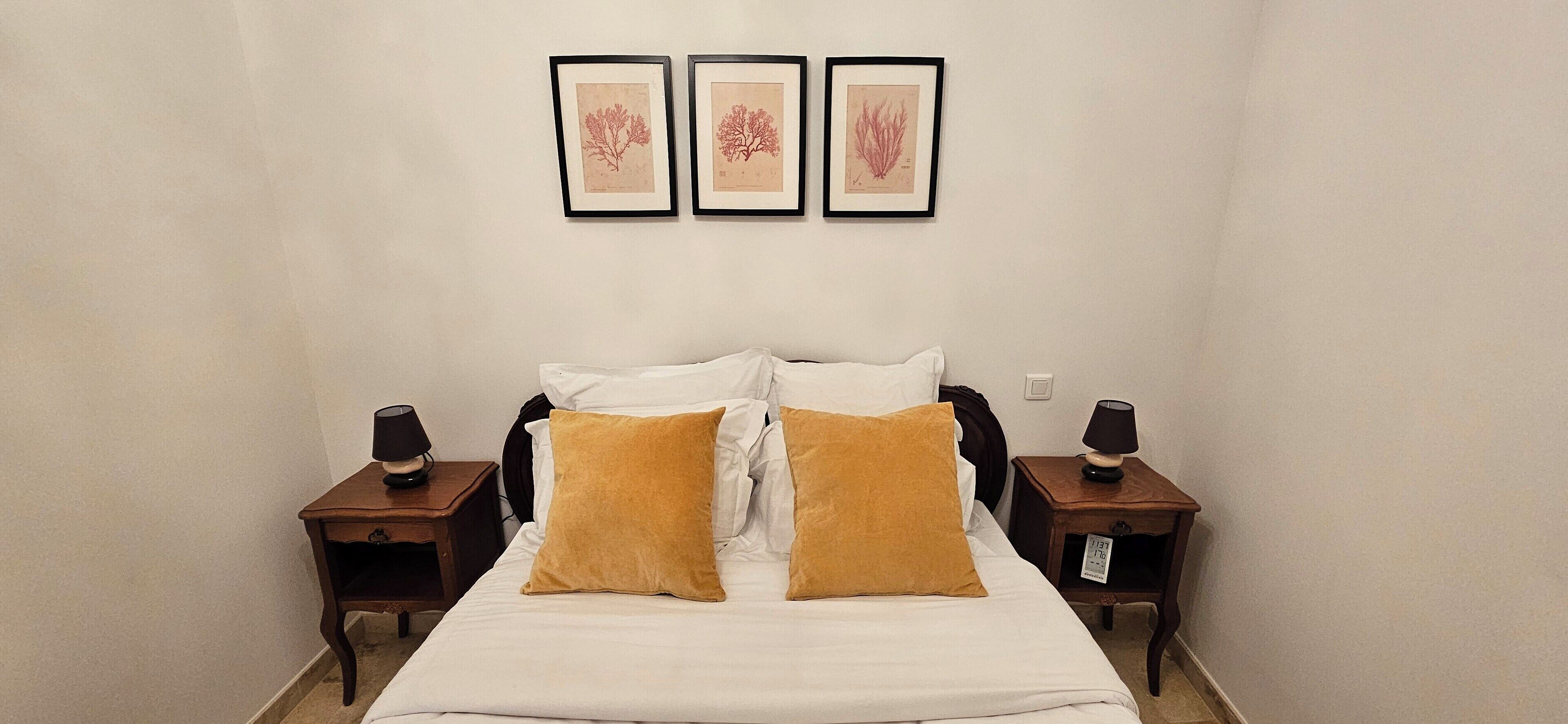 A neatly made bed with white linens and two mustard-colored pillows, flanked by two bedside tables with lamps. Three framed botanical prints adorn the wall above the bed.