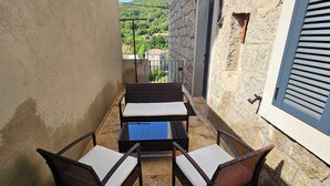 Un balcón exterior estrecho con una mesa y cuatro sillas, un muro de piedra y una ventana con contraventanas.