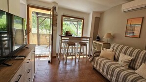 A cozy living area with a TV, a striped sofa, a dining table with chairs, and a view through a glass door. - Mini Caseddu T2, Pool & sea view, Alba Rossa N°11 (Serra-di-Ferro)