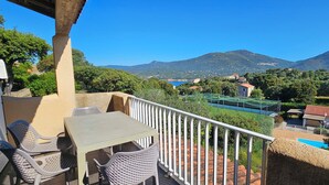 A balcony with a table and chairs, overlooking a mountainous landscape with a pool and tennis court. - T2 Dolce & Vita C5 sea and pool view (Propriano)