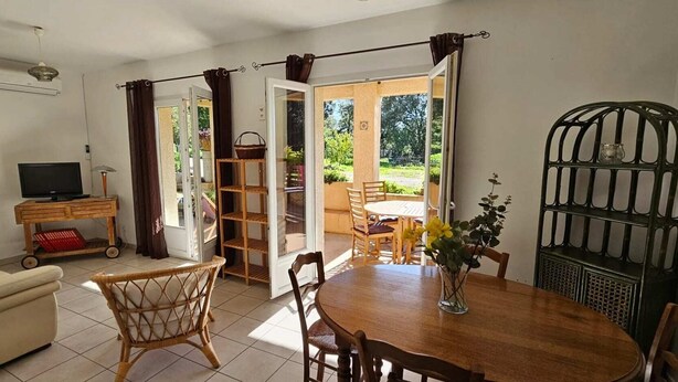A living room with a sofa, a TV, a wooden table with chairs, and a dining area with a table and chairs. There is a sliding glass door leading to an outdoor patio. - Peaceful T3 in Vigna Maggiore, Olmeto (Olmeto)