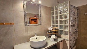 A bathroom with a round sink, a large mirror, a wooden shelf with toiletries, and a shower area with patterned curtains. - Peaceful T3 in Vigna Maggiore, Olmeto (Olmeto)