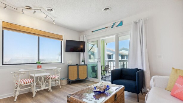 A room with a large window, a television, a dining table with chairs, a sofa, and a wicker coffee table.