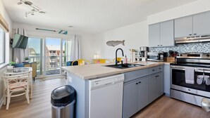 A modern kitchen with grey cabinets, a dishwasher, an oven, and a sink. There is a fish decoration on the wall, a window with curtains, and a small dining area with a chair and table.