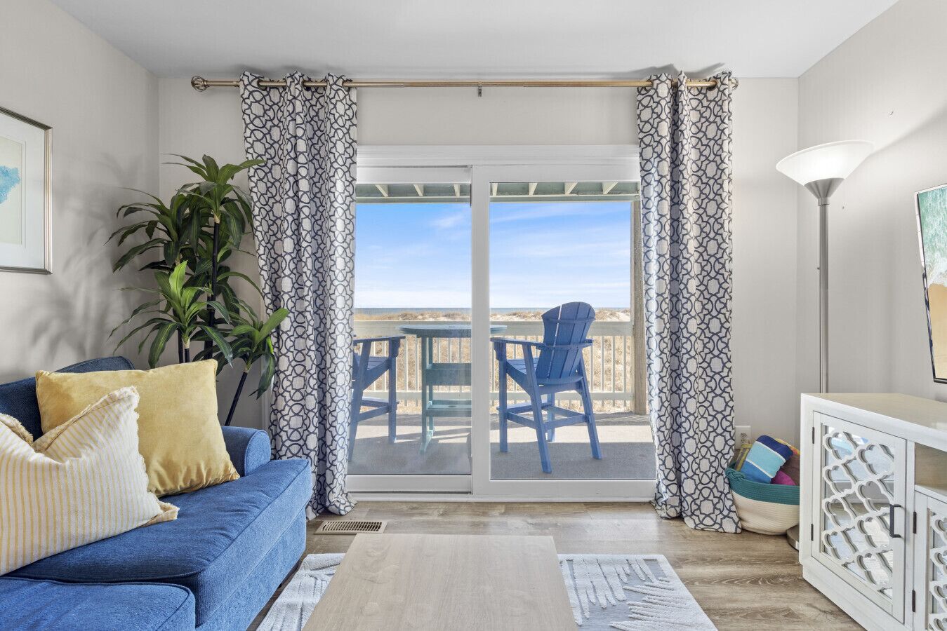 Una habitación con un sofá azul, una planta en maceta, una mesa con sillas y vista a la playa por un ventanal corredero.
