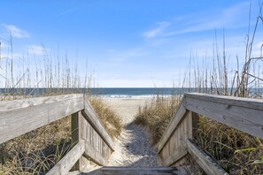 A wooden boardwalk leading to a beach with sand and sparse vegetation. - Remodeled Oceanfront Condo w/ Pool (Carolina Beach)