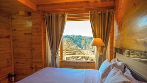 Habitación de cabaña de madera con cama, cortinas y ventana con vistas a una colina boscosa.