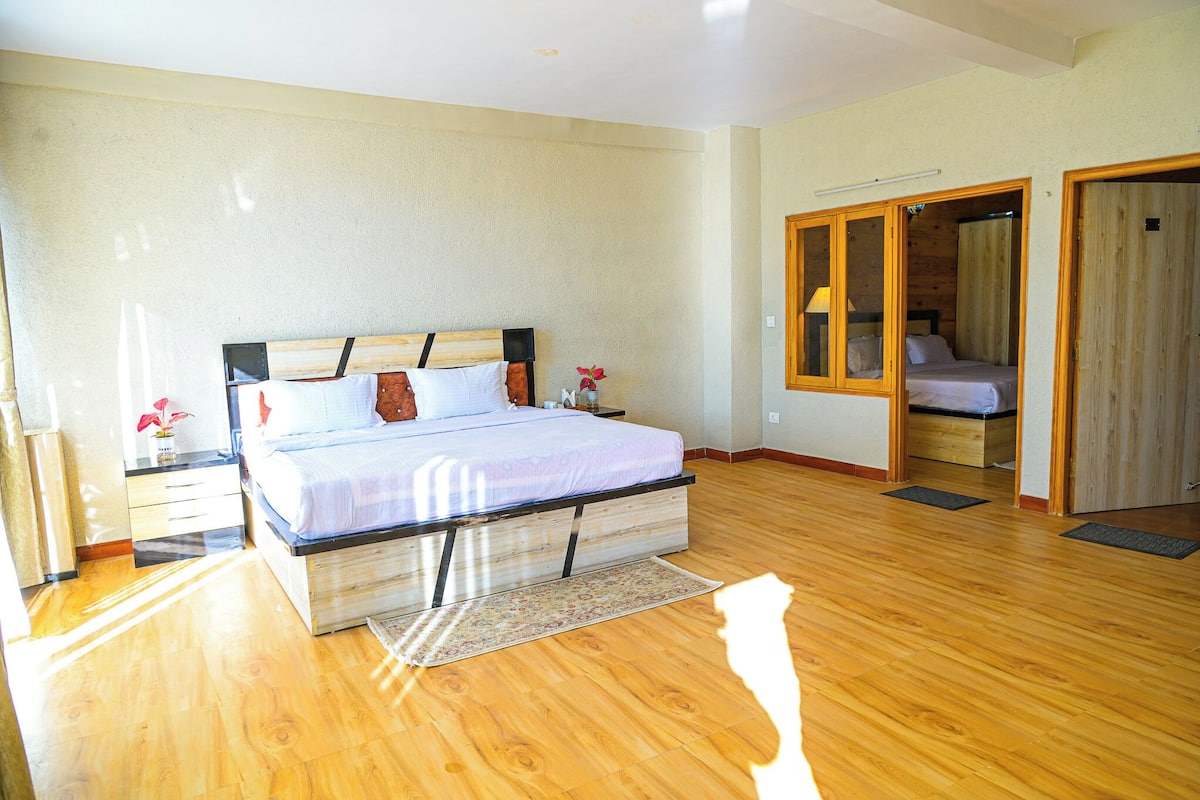 A bedroom with a wooden floor, a bed with a headboard, a nightstand, a mirror, and a door leading to another room.