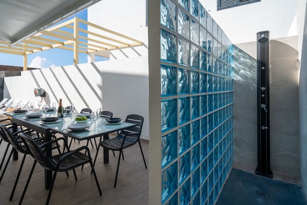 Comedor con mesa dispuesta para cuatro, pérgola en lo alto y pared de vidrio con cabina de ducha.