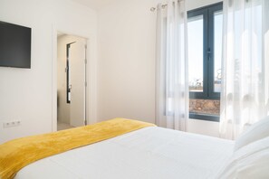 Un dormitorio con cama, ventana con cortinas, puerta y televisor instalado en la pared.