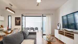 Una sala moderna con un sofá gris, una mesa de centro de vidrio, un televisor de pantalla plana y un comedor con una mesa y sillas de superficie de vidrio.